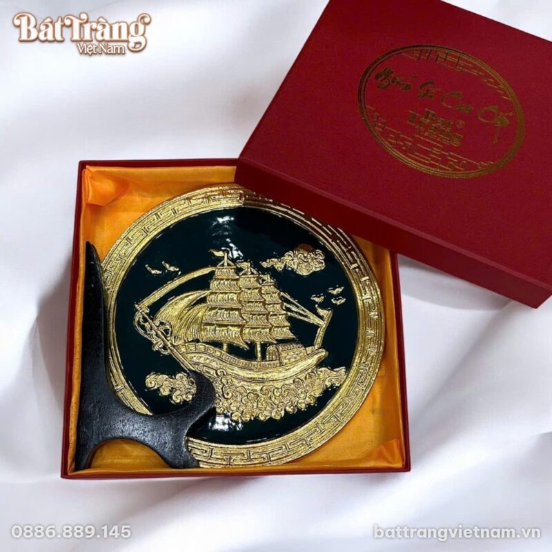 Đĩa gốm sứ trang trí hình tròn 'Thuận Buồm Xuôi Gió' với họa tiết thuyền buồm nổi 3D màu vàng kim rực rỡ trên nền men xanh ngọc lục bảo, viền hoa văn chữ vạn vàng kim, đặt trên giá đỡ gỗ đen. Sản phẩm nằm gọn trong hộp quà màu đỏ sang trọng có lót vải vàng cam, tất cả đặt trên nền vải trắng.